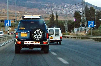 צילום מתוך רכב בדרך עירונית: רכב שטח כהה עם גלגל רזרבי נוסע לפנינו ורכב מסחרי לבן מימין. תמרורים כחולים משני צדי הכביש.