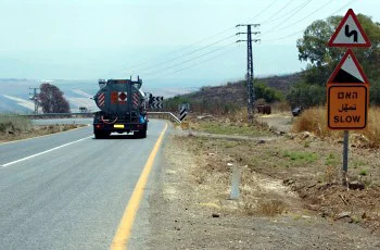 צילום מתוך רכב בדרך בין-עירונית: רכב כהה נוסע לפנינו, קו צהוב בשוליים, ומימין ניצבים תמרור משולש אדום ובו קו מתפתל ולוח כתום עם כיתוב.