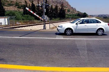 מפגש מסילת ברזל: מחסום מפוספס אדום-לבן מורם מעל הכביש, מכונית לבנה חולפת מתחתיו, וקו צהוב רחב בשולי הכביש.
