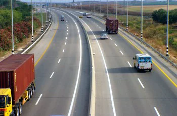 צילום מלמעלה של דרך רחבה: שלושה נתיבים לכל כיוון, משאיות עם מכולות בצד שמאל ורכבים נוספים בנתיבים, בין שדות פתוחים.
