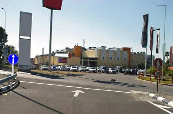 צילום מתוך רכב המתקרב לצומת: חץ לבן כלפי מעלה מסומן על האספלט, משמאל תמרור עגול כחול ובו חץ לבן כלפי מעלה, מימין תמרורים עגולים עם מסגרת אדומה, ומעבר לצומת מגרש חניה מלא מכוניות ומבנה מסחרי.