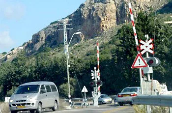 מפגש מסילת ברזל: מחסומים מפוספסים אדום-לבן במצב מורם, עמוד ובו משולש אדום, וסימן דמוי איקס אדום-לבן. רכבים עוברים במקום, וברקע צוק סלעי.