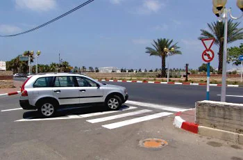 צילום בצומת עירוני: מכונית כסופה עומדת ליד פסים לבנים הצבועים לרוחב הכביש. על עמוד מימין משולש הפוך עם מסגרת אדומה, ומתחתיו תמרור עגול עם מסגרת אדומה ובו חץ שחור מתעקל וקו אדום. אי תנועה ואבני שפה אדום-לבן בצדדים.
