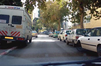 צילום מתוך רכב ברחוב עירוני: רכב מסחרי לבן עם פסים אדומים אלכסוניים נוסע לפנינו, ומכוניות חונות בשני צדי הכביש.