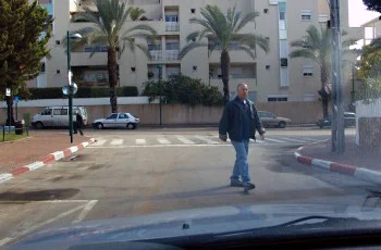 צילום מתוך רכב ברחוב עירוני: אדם במעיל כהה חוצה את הכביש לפנינו, ושפת המדרכה צבועה אדום-לבן. דקלים ומבני מגורים ברקע.