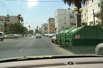 צילום מתוך רכב ברחוב עירוני: מכוניות חונות בשני צדי הכביש, מכולת אשפה ירוקה גדולה עומדת מימין, ומבנים ודקלים ברקע.