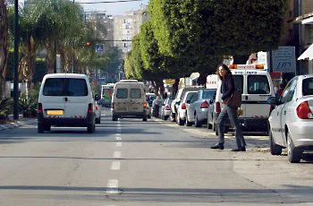 צילום מתוך רכב ברחוב עירוני: אדם חוצה את הכביש בין מכוניות חונות, ורכבים נוסעים לפנינו. עצים ומבנים ברקע.